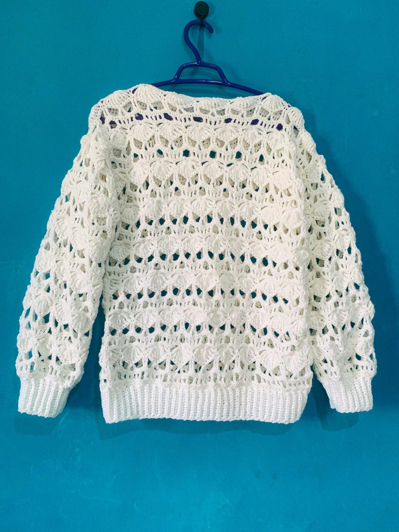 May include: A white, open-weave crochet sweater with long sleeves and a ribbed hem and cuffs. The sweater is hanging on a blue hanger against a blue background. The crochet pattern features a repeating shell design.