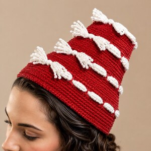 Peut inclure: Un bonnet en crochet rouge foncé orné de plusieurs rangées horizontales décorées de petites franges blanches. Le bonnet présente une forme structurée et légèrement conique, réalisé avec un fil dense et texturé de couleur rouge unie.