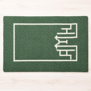 May include: A rectangular green crochet mat featuring a white border and a white maze-like pattern on the right side. The textured surface is composed of dense, uniform stitches, creating a consistent grid-like appearance throughout the piece.