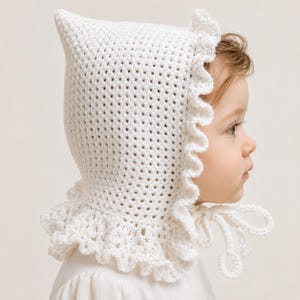 Peut inclure: Un bonnet en crochet blanc doté d'un sommet pointu, d'une bordure à volants délicate le long de l'ouverture du visage et du cou, et de deux fines attaches en crochet qui se nouent sous le menton. Le bonnet présente un motif en maille ajourée.