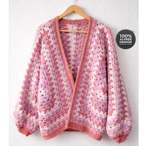 Könnte beinhalten: Handgefertigte Strickjacke mit offenem Schnitt und Granny-Square-Muster. Der Pullover hat eine Farbpalette aus Rosa, Lavendel und Weiß. Die Ärmel sind lang und voluminös. Der Text "100% AI-FREE DESIGN" ist auf einem runden Etikett zu sehen.