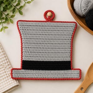 May include: A grey crochet pot holder, shaped like a top hat, with a black band and red edging. It features a hanging loop with a wooden bead. The pot holder is likely used in the kitchen.