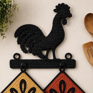May include: Black crochet rooster wall decor with a bar for hanging pot holders. Two triangular pot holders in yellow and orange with black floral designs are suspended below. Wooden utensils are visible in the background.