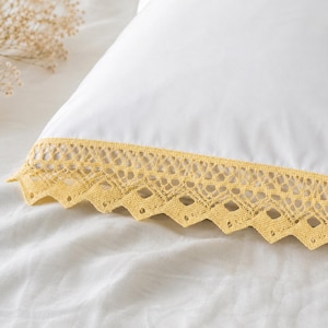 Pode incluir: Uma fronha branca com uma borda decorativa de renda de crochê amarela. O acabamento em renda apresenta um padrão aberto intrincado e uma borda em ziguezague com pequenos recortes circulares, contrastando com o tecido branco liso.