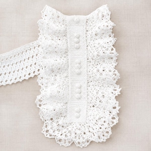 May include: A white crochet collar featuring a central vertical panel with six round buttons. The collar is adorned with intricate lace-like patterns and ruffled edges, extending into a wide, open-weave band on the left side of the piece.