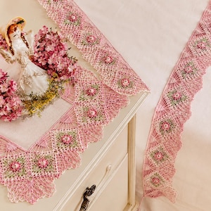 May include: A pink and white crocheted table runner featuring a repeating square motif with floral centers in shades of pink and green. The edges are finished with a delicate scalloped lace pattern. The runner is draped over a white surface.