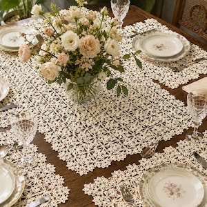 May include: A cream-colored crocheted tablecloth with a repeating floral pattern, featuring a central glass vase filled with white and pale pink roses, surrounded by matching place settings with floral-rimmed plates and clear crystal glassware.