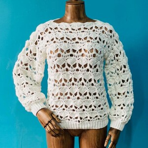 May include: A white, open-weave crochet sweater with long sleeves. The sweater features a scalloped neckline and cuffs, and a textured, shell-like pattern throughout the body. The garment is displayed on a wooden mannequin against a bright blue background.