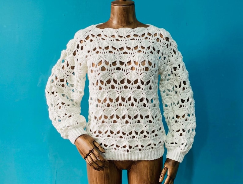 May include: A white, open-weave crochet sweater with long sleeves. The sweater features a scalloped neckline and cuffs, and a textured, shell-like pattern throughout the body. The garment is displayed on a wooden mannequin against a bright blue background.