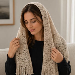 Può includere: Un copricapo all'uncinetto color beige caratterizzato da un intricato motivo traforato e delicate frange lungo i bordi inferiori. Il tessuto è leggero con una trama testurizzata e ariosa che ricade su testa e spalle, creando un aspetto morbido.