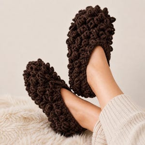 May include: A pair of dark brown, textured crochet slippers. The slippers are made with a thick yarn, creating a dense, nubby appearance. The feet are resting on a white, fluffy surface, and the person is wearing a cream-colored sweater.