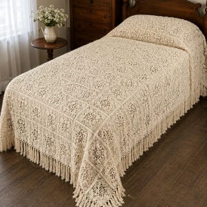 May include: A cream-colored bedspread featuring a repeating square crochet pattern with intricate floral motifs. The textile is finished with long, dangling fringe along the edges, covering the top and sides of a bed frame in a bedroom setting.