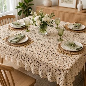 Puede incluir: Un mantel de ganchillo beige con un patrón floral circular repetitivo que cubre una mesa de comedor. La mesa está puesta con cuatro platos blancos sobre manteles individuales redondos de mimbre, con servilletas verde salvia y flores blancas.