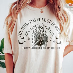 May include: Cream-colored t-shirt with a black graphic featuring a skeleton reading a book, surrounded by stars, bats, and the text "THE WORLD IS FULL OF HORROR" and "THROW ANOTHER BOOK ON THE TBR".