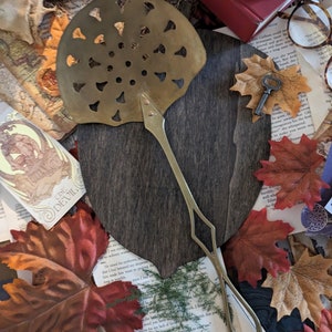 May include: A brass, decorative, fan-shaped,  kitchen utensil with a long handle. The utensil is on a dark wood surface with a key and fall leaves around it.