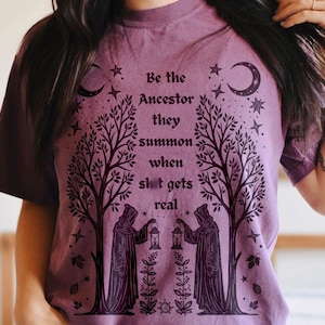 May include: A mauve-colored t-shirt with a graphic design featuring two robed figures holding lanterns beneath stylized trees. The text reads "Be the Ancestor they summon when sh!t gets real" with crescent moons and stars.