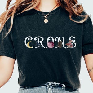 May include: Dark gray t-shirt with the word "CRONS" in white, featuring a crescent moon, raven, cauldron, bundle, and mushroom graphic. The shirt is made of a soft material.
