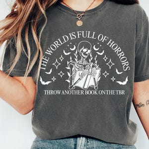 May include: Dark gray t-shirt with a white graphic of a skeleton reading a book, surrounded by bats, moons, and stars. Text reads "THE WORLD IS FULL OF HORRORS" and "THROW ANOTHER BOOK ON THE TBR".