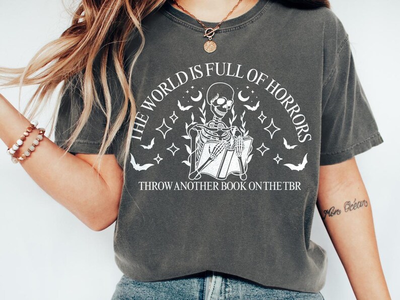 May include: Dark gray t-shirt with a white graphic of a skeleton reading a book, surrounded by bats, moons, and stars. Text reads "THE WORLD IS FULL OF HORRORS" and "THROW ANOTHER BOOK ON THE TBR".
