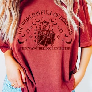 May include: A maroon t-shirt with a graphic featuring a skeleton reading a book, surrounded by bats, stars, and the text "THE WORLD IS FULL OF HORRORS" and "THROW ANOTHER BOOK ON THE TBR".