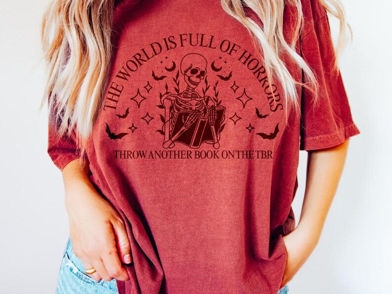 May include: A maroon t-shirt with a graphic featuring a skeleton reading a book, surrounded by bats, stars, and the text "THE WORLD IS FULL OF HORRORS" and "THROW ANOTHER BOOK ON THE TBR".