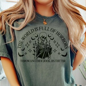 May include: A faded green t-shirt with a black graphic of a skeleton reading a book, surrounded by bats, stars, and the text "THE WORLD IS FULL OF HORRORS" and "THROW ANOTHER BOOK ON THE TBR".