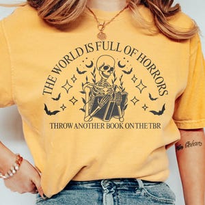 May include: Mustard yellow t-shirt with a black graphic of a skeleton reading a book, surrounded by bats, stars, and moons. Text reads "THE WORLD IS FULL OF HORRORS" and "THROW ANOTHER BOOK ON THE TBR".