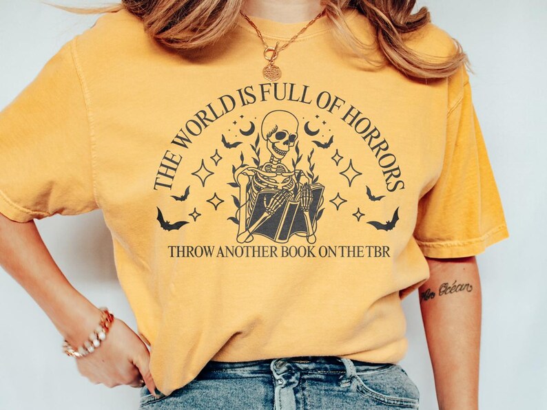 May include: Mustard yellow t-shirt with a black graphic of a skeleton reading a book, surrounded by bats, stars, and moons. Text reads "THE WORLD IS FULL OF HORRORS" and "THROW ANOTHER BOOK ON THE TBR".