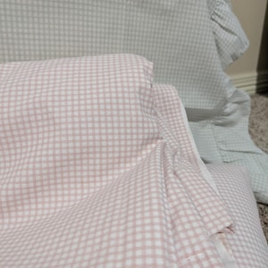 May include: A folded textile featuring a light pink and white gingham check pattern. In the background, a separate piece of fabric with a light green and white gingham check pattern and a ruffled edge is partially visible.