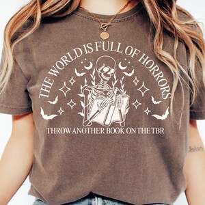 May include: Brown t-shirt with a white graphic of a skeleton reading a book, surrounded by bats, moons, and stars. Text reads "THE WORLD IS FULL OF HORRORS" and "THROW ANOTHER BOOK ON THE TBR".