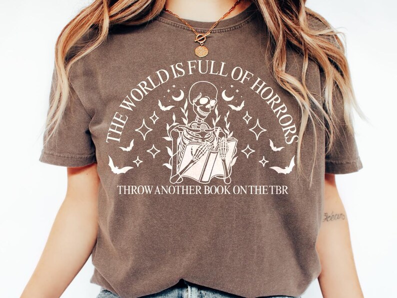 May include: Brown t-shirt with a white graphic of a skeleton reading a book, surrounded by bats, moons, and stars. Text reads "THE WORLD IS FULL OF HORRORS" and "THROW ANOTHER BOOK ON THE TBR".
