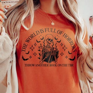 May include: Orange t-shirt with a skeleton graphic holding a book, surrounded by bats, stars, and moons. The text reads "THE WORLD IS FULL OF HORRORS" and "THROW ANOTHER BOOK ON THE TBR".