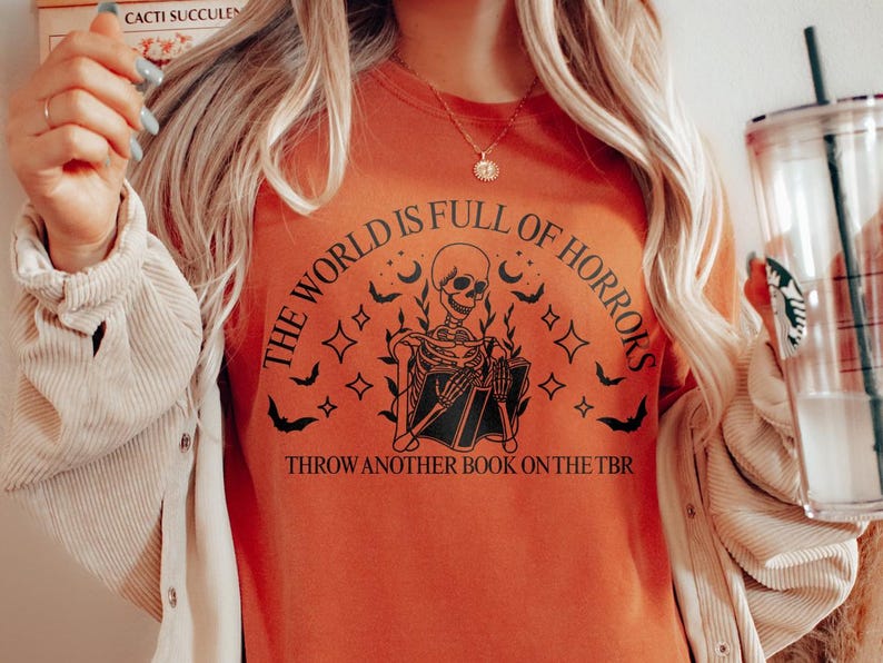 May include: Orange t-shirt with a skeleton graphic holding a book, surrounded by bats, stars, and moons. The text reads "THE WORLD IS FULL OF HORRORS" and "THROW ANOTHER BOOK ON THE TBR".