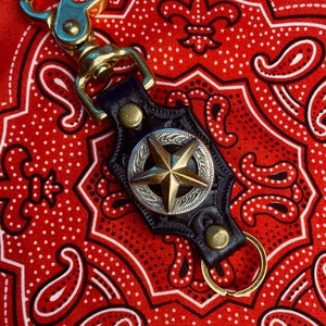 May include: A black leather keychain with a brass star and a silver-colored decorative border. The keychain has a brass clasp and a brass key ring. The background is a red bandana with a white paisley pattern.
