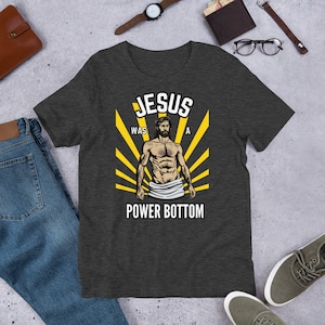 May include: A dark heather grey t-shirt with a graphic of a muscular Jesus figure with the text "Jesus was a power bottom".