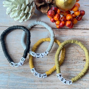 Puede incluir: Tres pulseras de cuentas con cuentas de letras blancas que deletrean las palabras "ASAP", "PUSH" y "HWLF". Las pulseras están sobre una superficie de madera con una piña, un adorno dorado y bayas rojas.