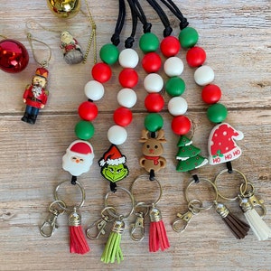 Puede incluir: Cinco llaveros con temática navideña con cuentas rojas, verdes y blancas. Cada llavero presenta un amuleto de personaje navideño diferente, que incluye a Papá Noel, el Grinch, un reno, un árbol de Navidad y un gorro de Papá Noel. Los llaveros tienen anillas de llaves plateadas y borlas en rojo, verde y marrón.