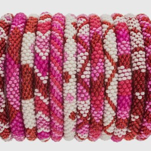 Puede incluir: Un conjunto de pulseras de cuentas en tonos rojos, rosas y blancos. Las pulseras están hechas de pequeñas cuentas redondas y presentan una variedad de patrones, incluyendo rayas y zigzags. Las pulseras están apiladas, creando un accesorio colorido y texturizado.