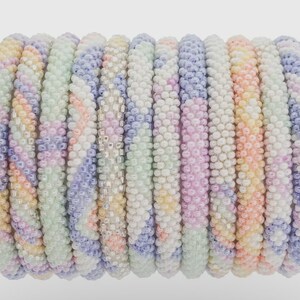 Puede incluir: Una pila de pulseras de cuentas coloridas en tonos pastel de rosa, morado, verde y amarillo. Las pulseras están hechas de pequeñas cuentas redondas y tienen un patrón de chevron.
