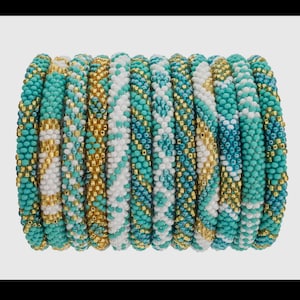 Puede incluir: Una pila de pulseras de cuentas coloridas en tonos turquesa, blanco y dorado. Las pulseras están hechas de cuentas pequeñas, estrechamente tejidas y tienen un patrón geométrico.