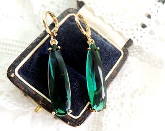 May include: A pair of gold-toned leverback earrings with large, emerald-green teardrop-shaped pendants. The earrings are displayed in a vintage jewelry box with a dark blue velvet interior, set against a backdrop of white lace.