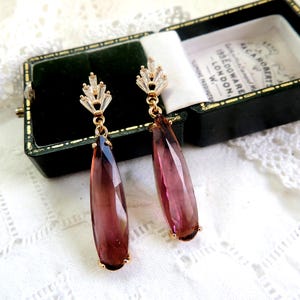 Può includere: Un paio di orecchini pendenti con una pietra preziosa a forma di lacrima di colore viola e rosa e un gruppo di strass trasparenti nella parte superiore. Gli orecchini sono in una piccola scatola verde con il testo "ISLEDGWARE LONDON W." sul coperchio.