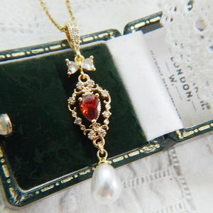 May include: A gold-tone necklace pendant featuring a pear-shaped red gemstone set in an ornate filigree frame with small clear crystals. A small bow-shaped crystal accent sits above the frame, and a white teardrop pearl hangs at the bottom.
