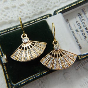 Peut inclure: Une paire de boucles d'oreilles pendantes dorées en forme d'éventails ouverts. Chaque boucle présente une pierre ronde transparente au sommet et plusieurs rangées de petites pierres scintillantes incrustées dans la structure métallique.