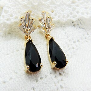 May include: A pair of gold-coloured drop earrings featuring black, teardrop-shaped gems. The earrings have a cluster of clear, baguette-cut crystals at the top. The earrings are set against a white, lace-like background.