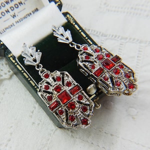 Könnte beinhalten: Ein Paar silberfarbene Ohrringe im Art-Deco-Stil mit roten Edelsteinen. Jeder Ohrring hat ein rechteckiges Design mit einem zentralen quadratischen Edelstein und kleineren Edelsteinen. Die Ohrringe werden in einer Vintage-Schmuckschatulle präsentiert.