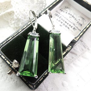 Op de afbeelding: Zilverkleurige oorbellen met grote, rechthoekige, groene edelstenen. De oorbellen worden gepresenteerd in een vintage zwarte sieradendoos met een witte binnenkant. De edelstenen zijn facetgeslepen.