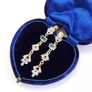 May include: A pair of dangle earrings with light blue rectangular stones and clear crystal accents. The earrings are in a blue velvet heart-shaped jewelry box.