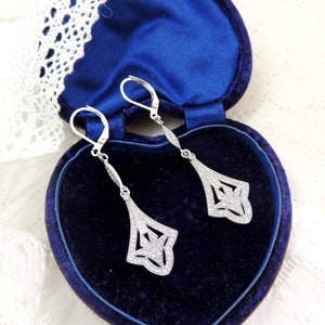 May include: A pair of silver dangle earrings with a geometric design, set with clear cubic zirconia stones. The earrings are displayed in a blue velvet heart-shaped jewellery box.