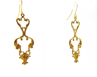 May include: A pair of gold-colored filigree earrings. Each earring features an ornate, openwork design with a central heart-shaped element and decorative flourishes. The earrings have a hook closure and are set against a white background.